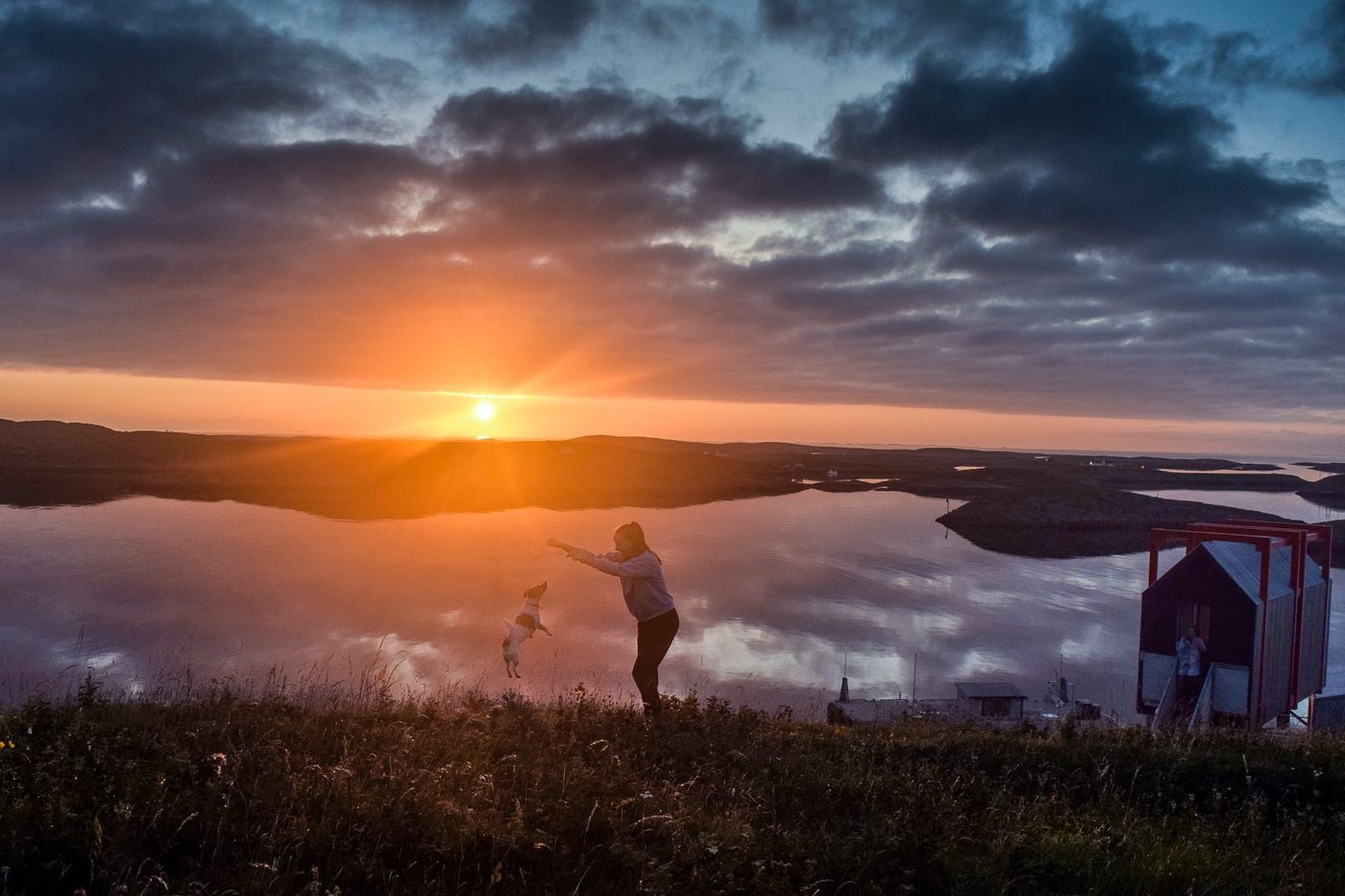 Andrea playing with her dog Leo on a hillside above the fjord at Fleinvær, Norway, at sunset, with a small red cabin nearby.
