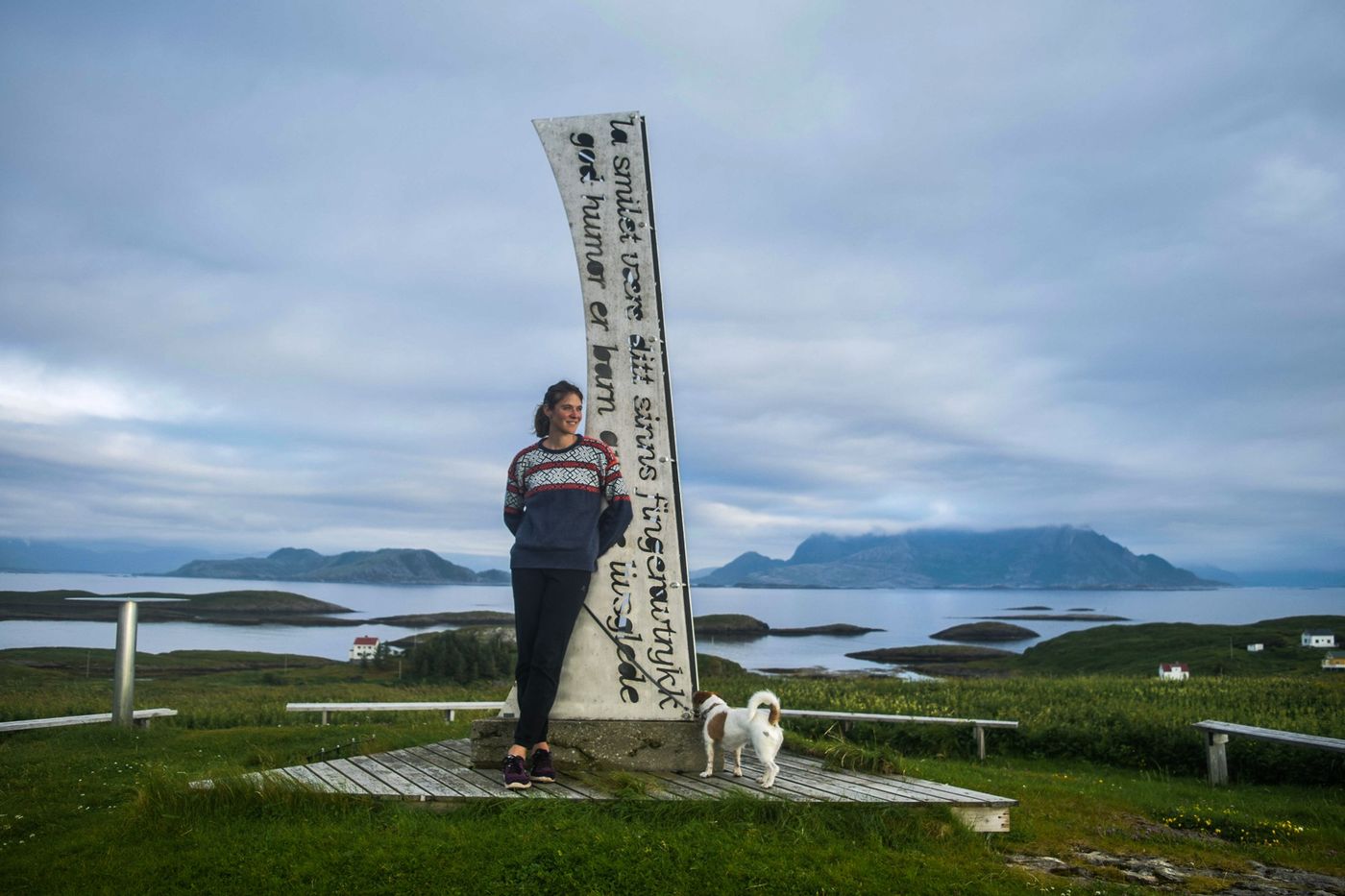 Andrea standing beside a tall wooden sail sculpture overlooking the fjord and islands at Fleinvær, Norway, with her dog Leo nearby.