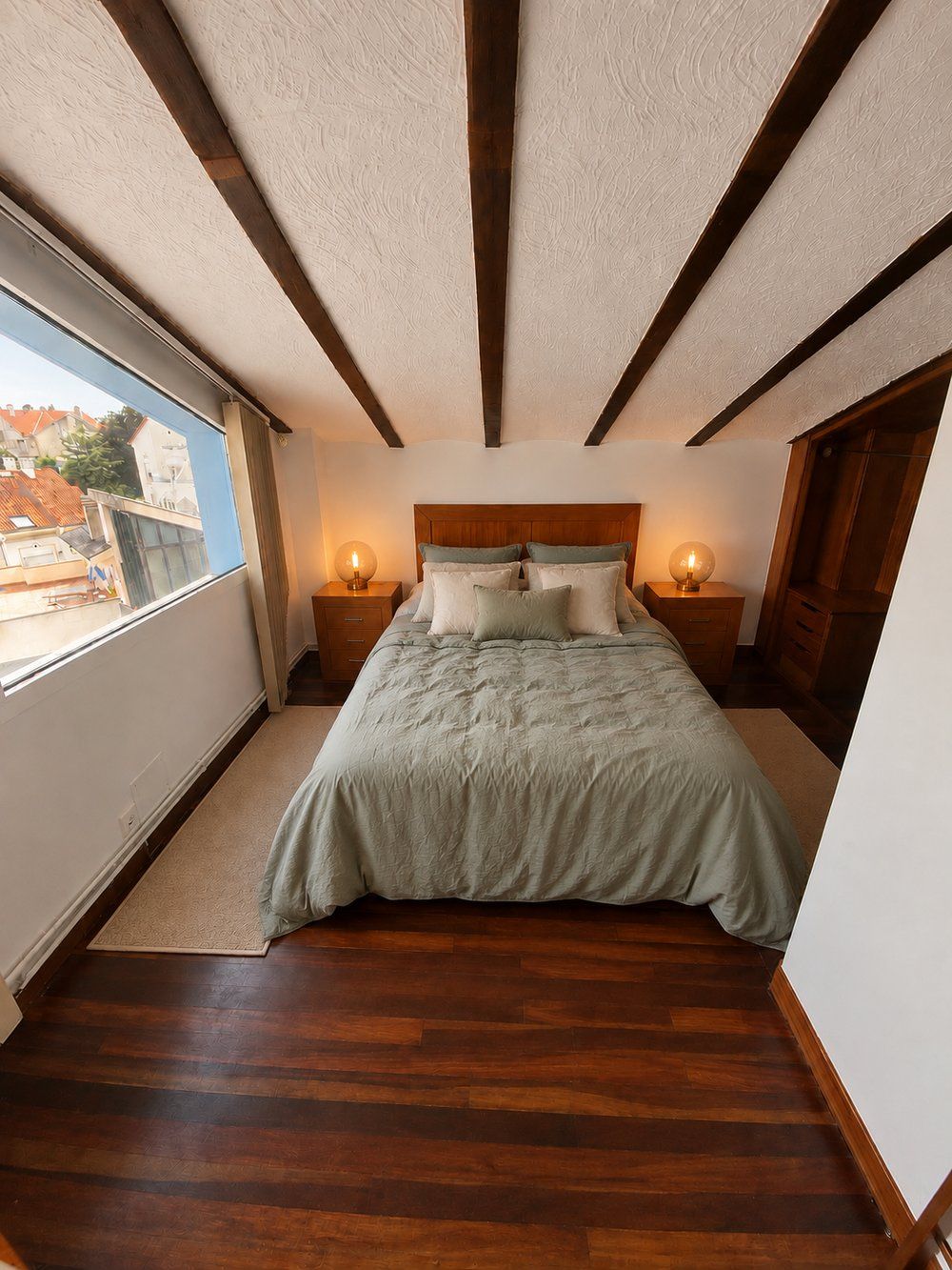 Un dormitorio abuhardillado con techos de vigas, ropa de cama verde salvia y una ventana con vistas a los tejados.