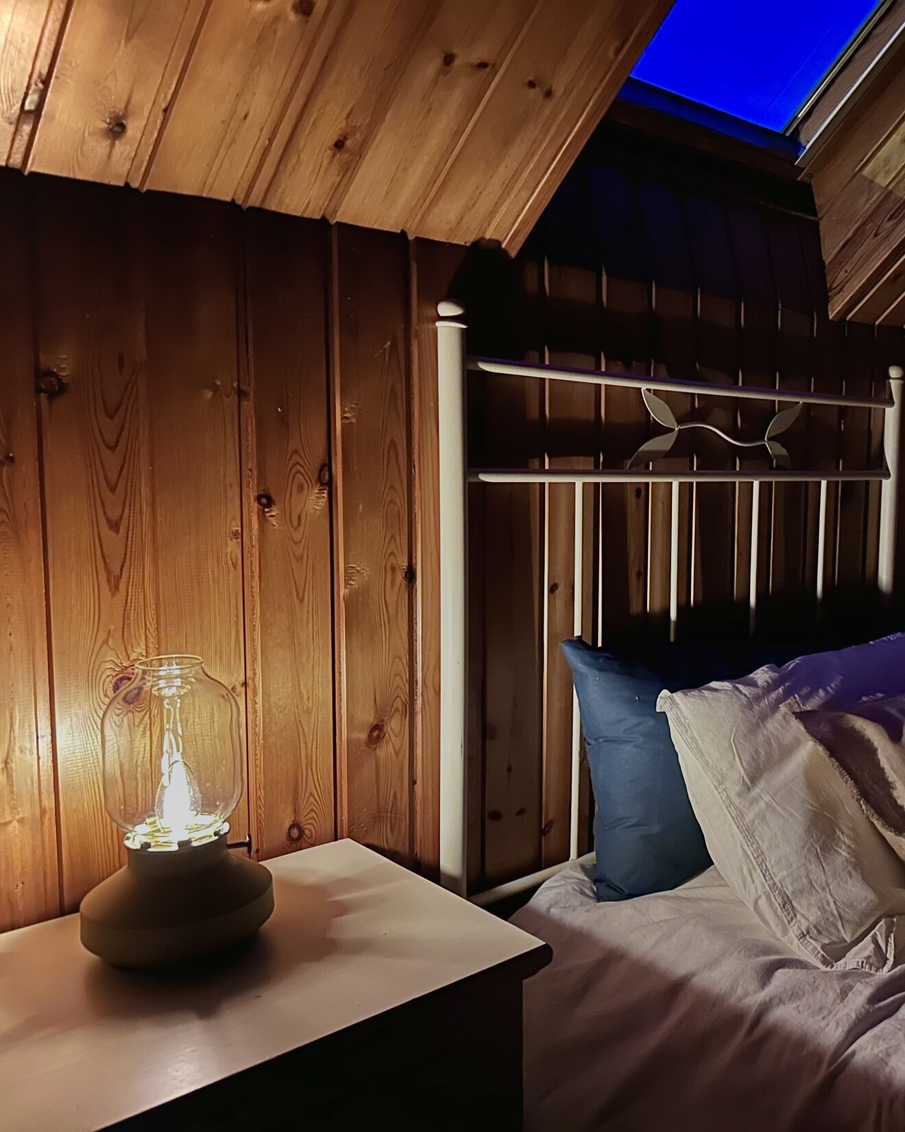 A lit lantern-style lamp on a bedside table in a wood-panelled attic room, with blue dawn light through the skylight.