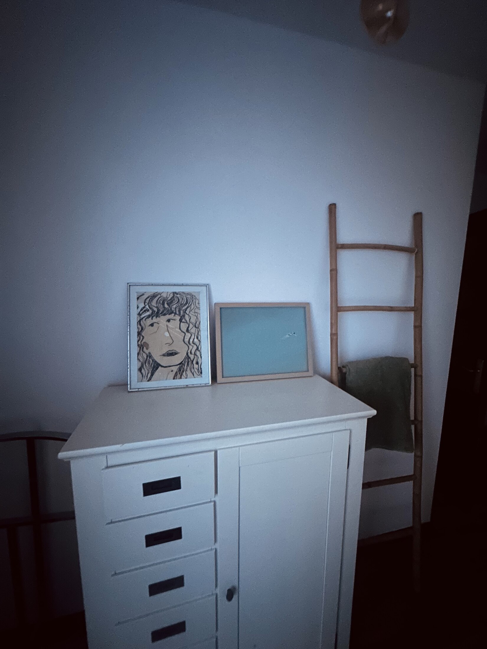 Framed artwork leaning on a white dresser, beside a bamboo ladder draped with a towel.