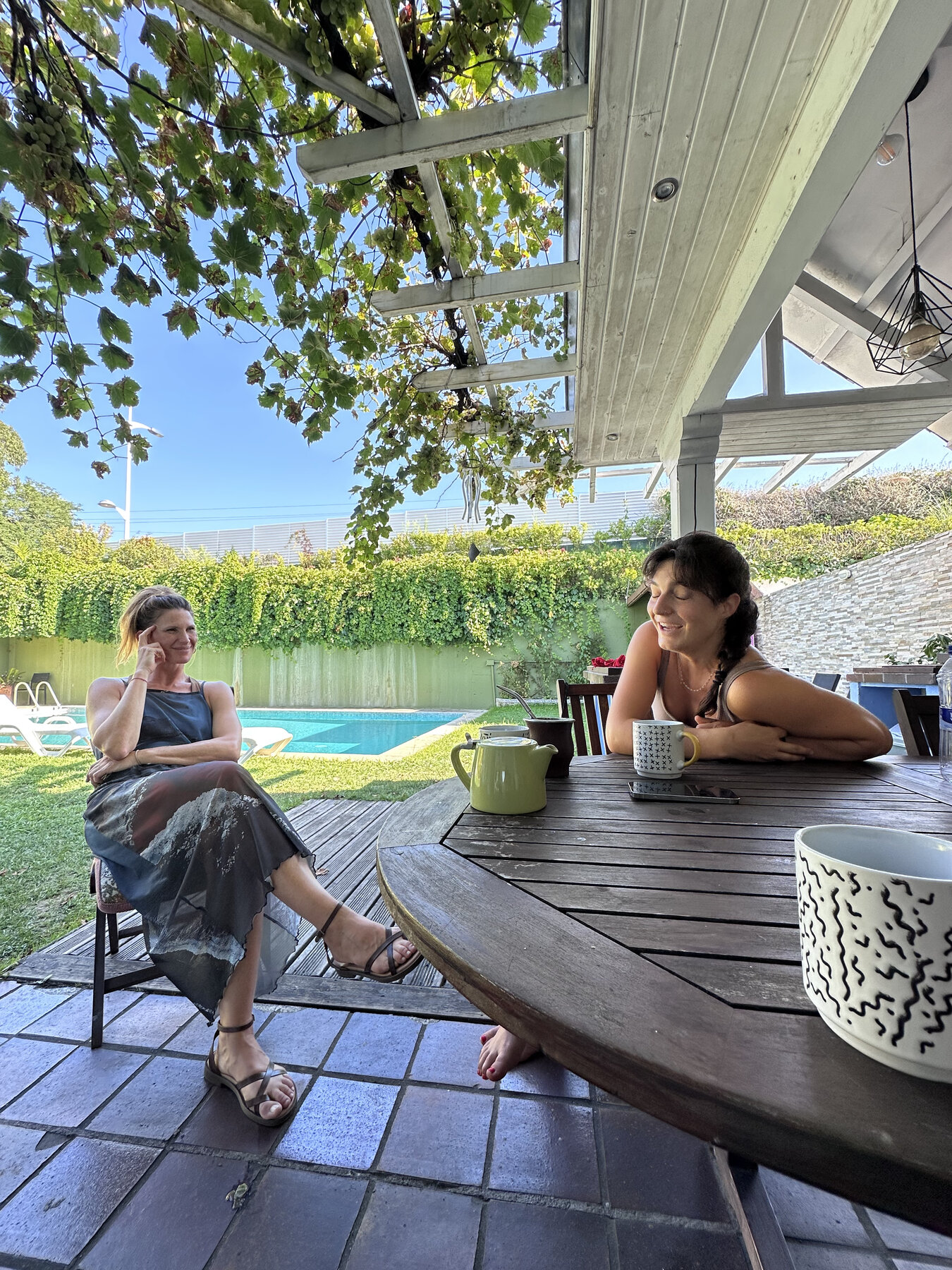 Two guests sharing coffee at the outdoor table under the pergola, with the pool behind them.