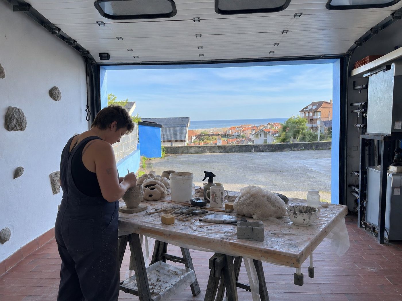 An artist working at a clay-covered table in a garage studio open to a sea view, shaping a piece of pottery.