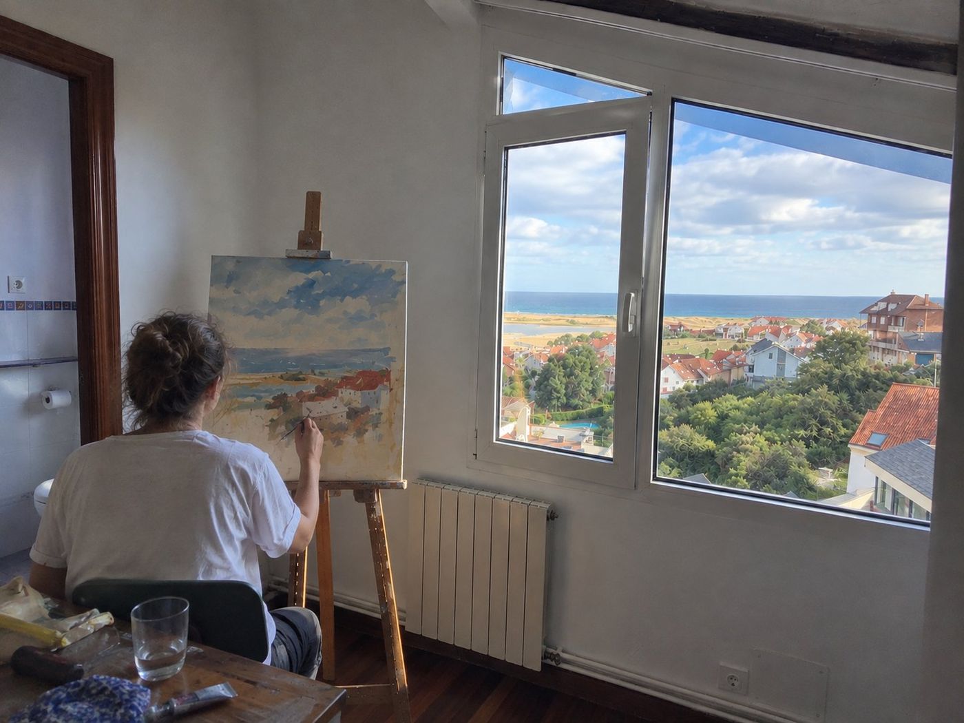 An artist painting at an easel in a bright room overlooking the coast, with painting supplies on a nearby table.